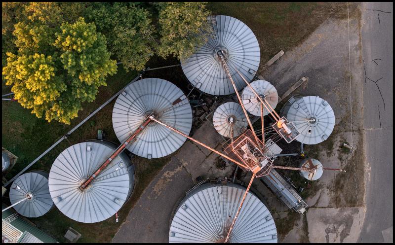 Looking straight down on a group of about 9 metal silos of various diameters, connected on top by thin metal pipes that appear somewhat corroded. 