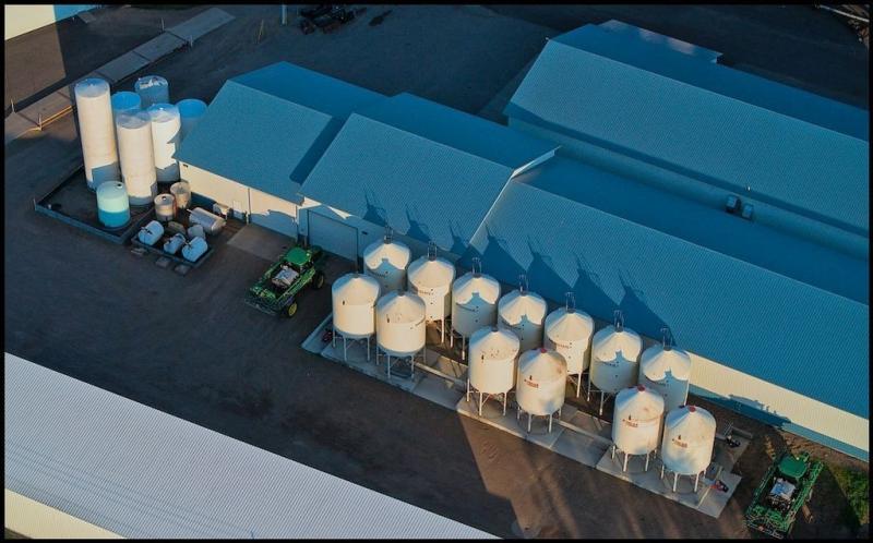 Aerial view of some very tidy metal storage structures with blue roofs and white walls. On one side of builders are a bunch of very large white tanks. A green tractor is parked near a garage type door.