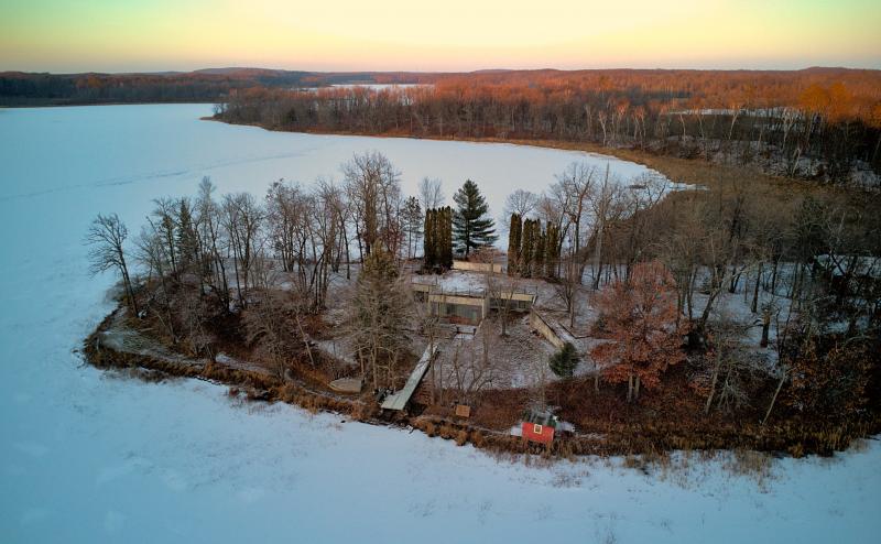 Aerial view of an underground cabin, built into a small hill on a small peninsula on a frozen lake. 