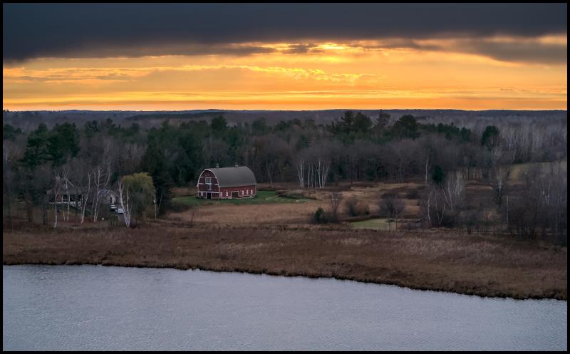 Aerial view of an upkept red barn with white trim. In forground is a lake and the sun is setting. The sky is a warm orange as the sun peeks through an overcast sky shortly before dusk.