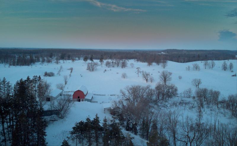 Drone photo of farm yard and red barn . Fields are covered with snow. Sky is blue with wisps of clouds.