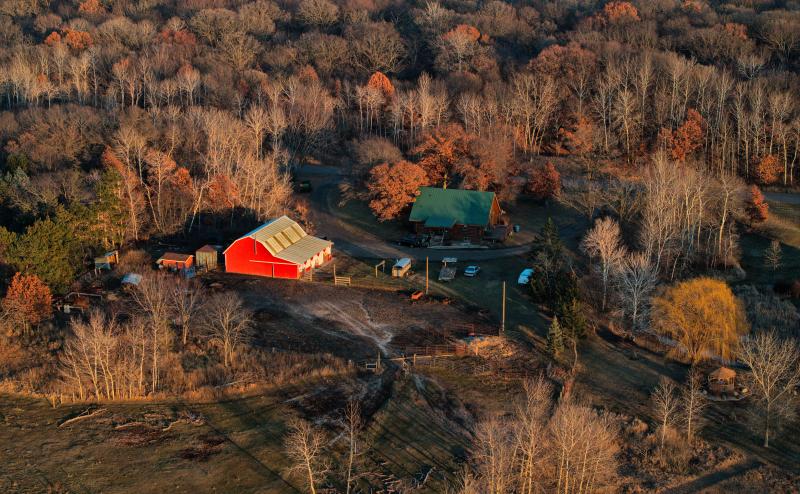 Aerial photo of a red barn and brown log house in a forest of mostly bare trees. Many of the trees are catching light and casting long shadows from a setting sun. 