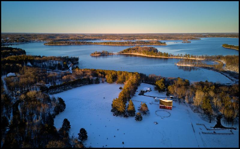 In the foreground we have a red barn and small corral. Fields are covered in snow. In background, we have a large lake with many islands and penninsulas. The water is mostly calm and open, but hints of ice along the edges are visible.