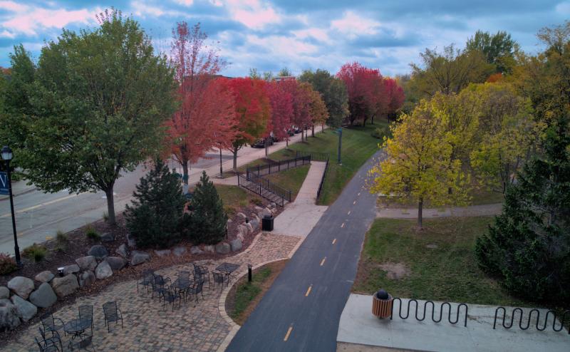 A bike path across divides the image with trees on either side. On the left side, many of the trees are various shades of red and in the foreground is a patio with metal patio chairs and tables. 
