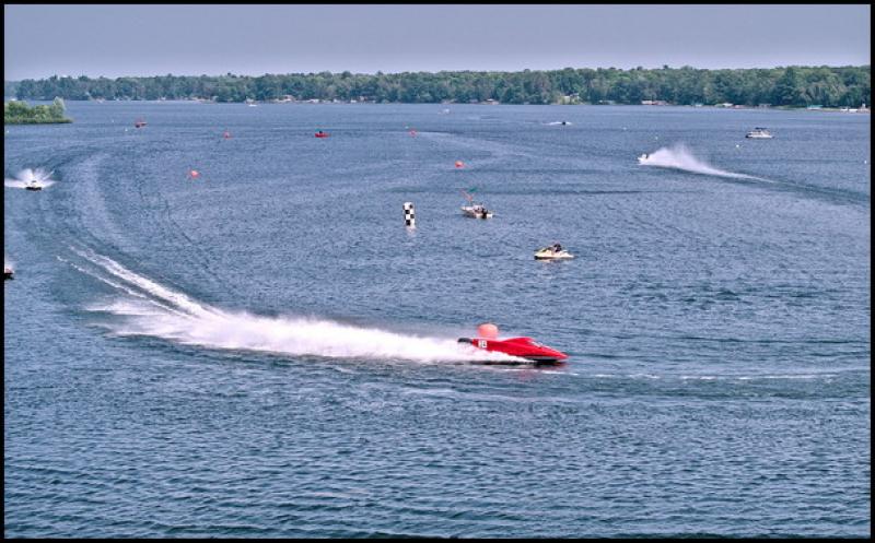 In the foreground red powerboat is make a turn around a large orange buoy. In the background, you can see other boats, at least two of which are part of the race and creating a large wake. 