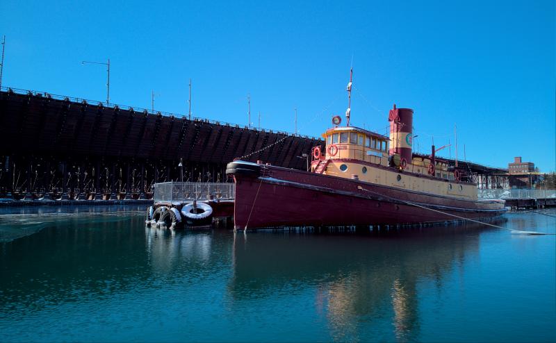 Old read and yellow tugboat tied off at dock in front of large ore dock for loading Great Lakes ore ships. Water is open around the boat, but ice is visible to the left side of the image.