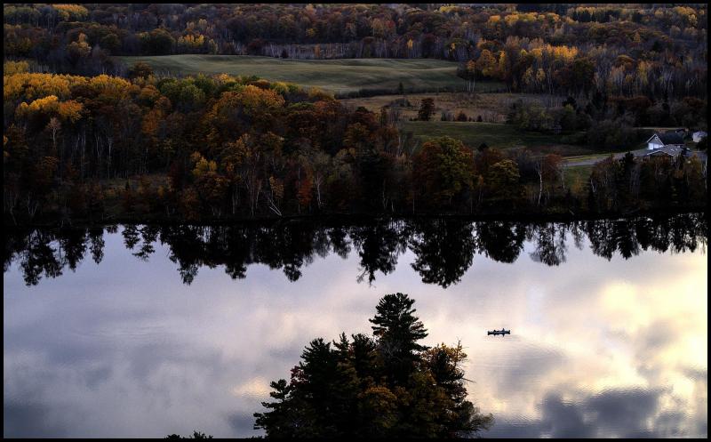 Aerial view of calm lake with canoe. Colorful autumn trees on the shore. Rolling fields in background.