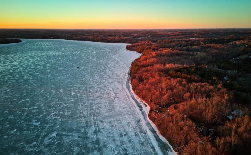 An aerial view of a frozen lake near sunset. We catch a little color in the sky and in the trees along the shore. A solitary ice fishing house stands on the frozen lake.