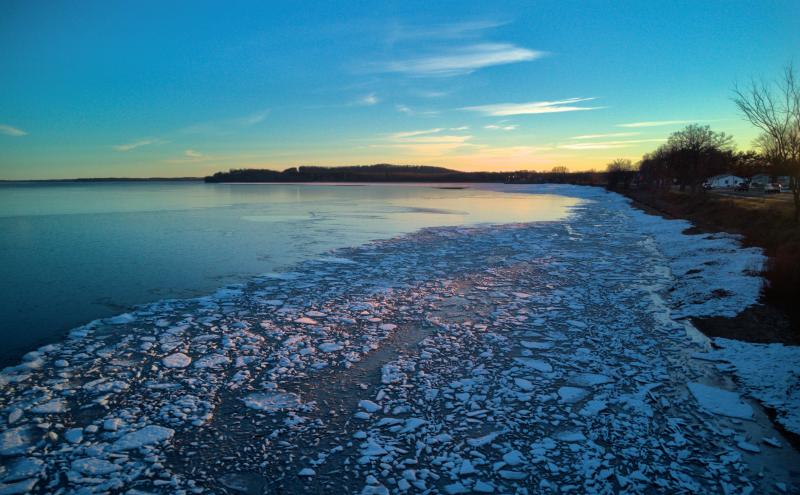 Looking along the shore of a large lake. A strip of previously broken ice has frozen back together and further out we see smooth ice and then open water. The sky is blue. 