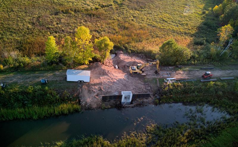 Aerial view of a backhoe parked at a site where work is happening on culvert/dam between two swamps. Also in picture is a small red bobcat type tool and box trailer to store equipment. 
