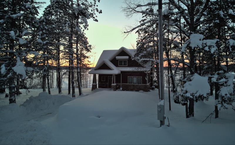 Snow covered house nestled between groves of pine trees. Frozen lake in background with hints of sunset peeping through.