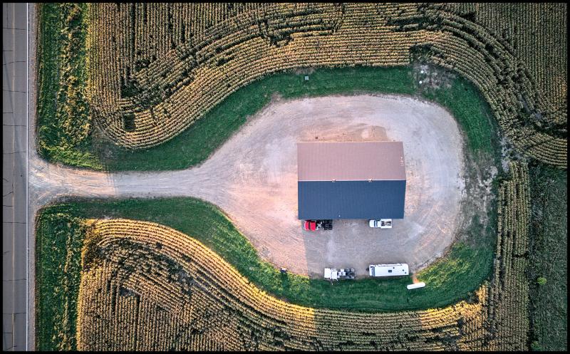 Looking straight down at a small building in the middle of an oval  parking lot, surrounded by farm fields that wrap around the parking lot. A gravel road leads out to the main road.