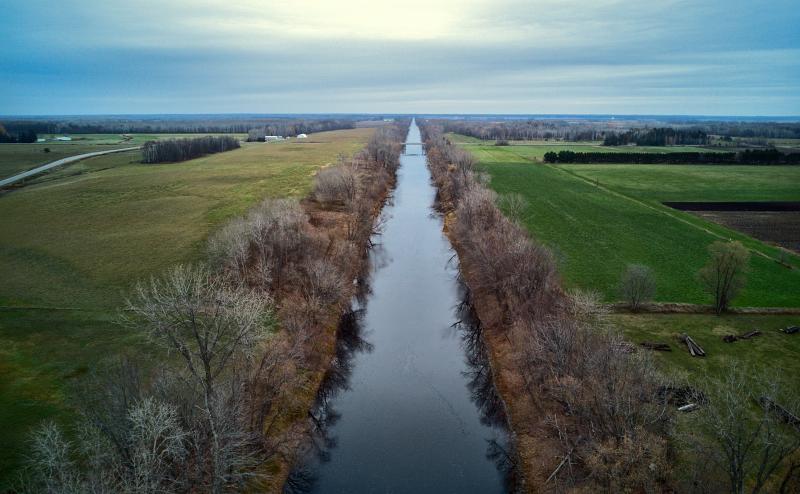 A straight channel of water bisects the image from top to bottom. A line of trees border the banks on either side and then farm fields. In the distance we can see a forest on either side of the channel. It's a cloudy day.