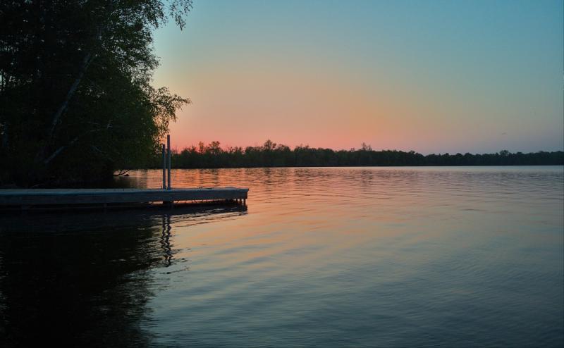 Looking across the end of a dock with tree lined shore on left and orange reflection of sunset in the background. The water is fairly still with mild ripples. 