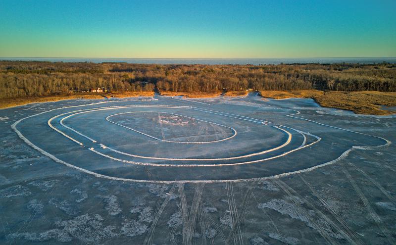 Aerial photo of a frozen lake with the borders of a race track track outlined in snow. There is no one on the lake or on the race track. It's emply. Beyond the lake is a forest of bare trees and in the far background another much larger lake. 