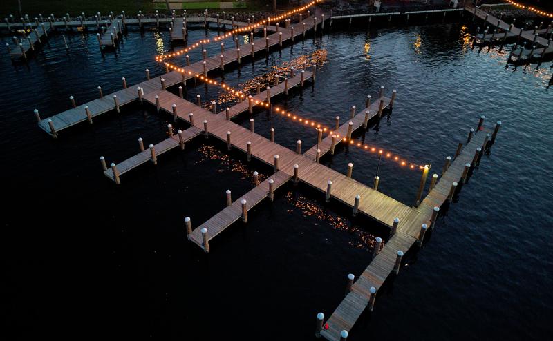 Looking down at an angle at a dock with three empty boat slips on either side. It's getting dark and the dock is strung with small white light bulbs that reflect off the water and the wooden dock. The water is calm.