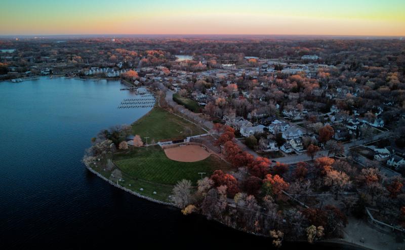 Looking down at neighborhood of mostly single family homes along side a large lake. A baseball field lies between the lake and many of the houses. The business part of town is vaguely visible in the distance. Sky indicates it is close to sunset.