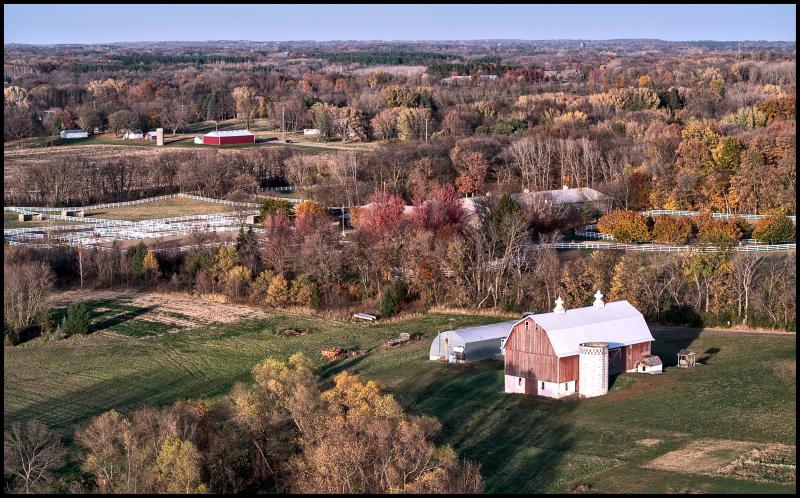 Aerial view of a faded red barn. In the background we can see a field divided up by many white fences. Most of the leaves have fallen, but a few trees hold onto faded leaves.