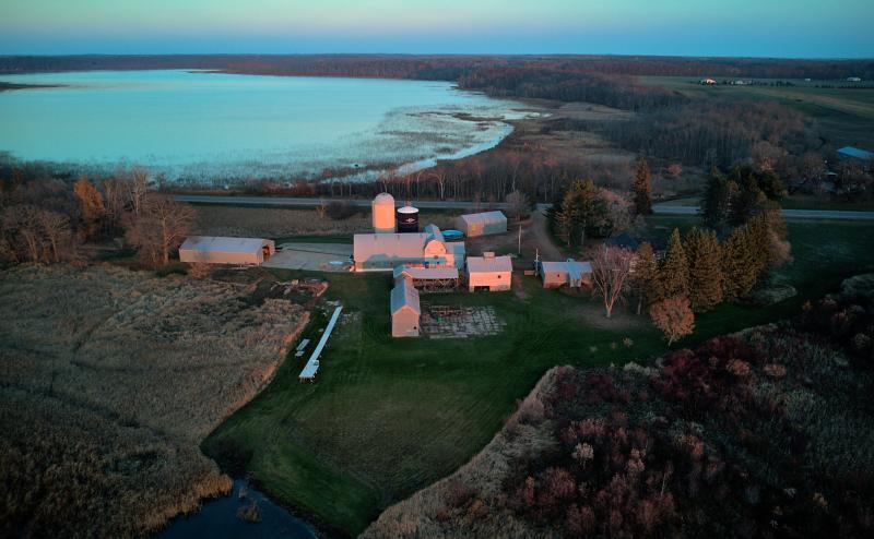 Aerial view of a farm house, barn, and other buildings situated between two lakes. A road passes by on the far side of the farm, the freshly cut lawn is bordered by brown tall grass. The buildings are tinted pink from the sunset happening behind us. 