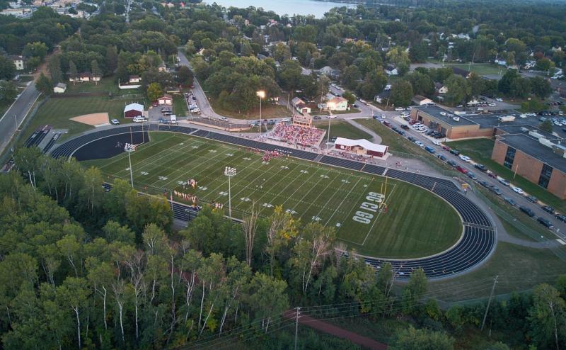 Aerial view of a high school football field with several hundred fans in the bleachers. Woods in foreground and a lake in the background. Field lights are on, but it's still daylight.