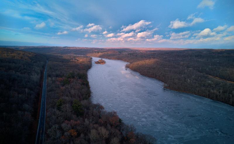 Aerial view of frozen river. The sky is blue and there are gentle rolling hills with forests on either side of the river. A two lane highway runs along the left side of the river. The sky is blue with scattered clouds.