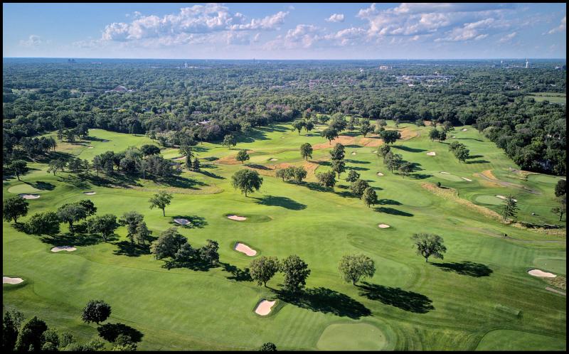 Aerial view of a golf course with many small sand traps. Water towers and other scattered low rise buildings pertrude about the tree line in the background. Sky is blue with scattered clouds.