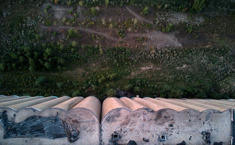 Looking straight down the side of a large abandoned grain elevator. We can see some graffitti on the roof and the curved edges of the side by side silos.