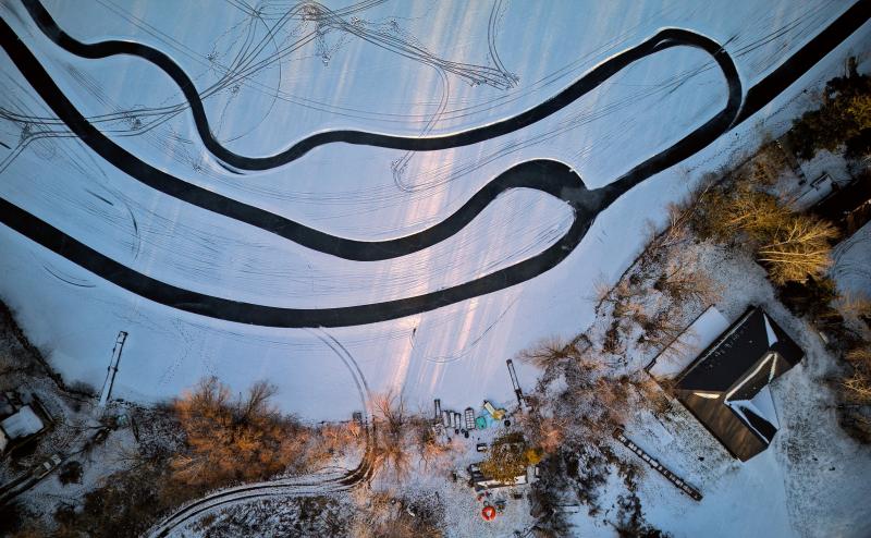 Looking straight down on frozen lakeshore. Buildings and docks are visible on shore, but focus of photo is a series of paths plowed in the snow on the ice that seem to be several windy paths for ice skating that intersect. We only see portions of the paths. 