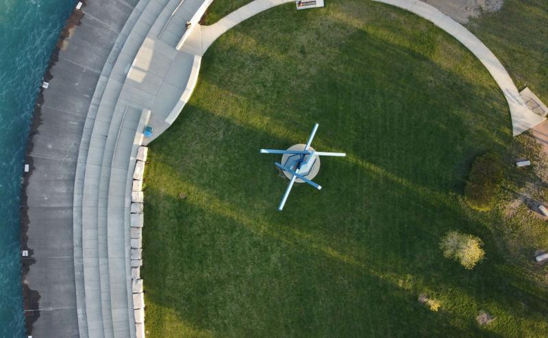 Arieal photo looking straight down on scupture along Lake Michigan. Statue is blue and look a bit like a very abstract windmill. Green is very green around statue and left side of image includes walkway and edge of water. 