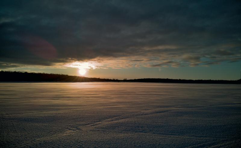 Just before sunset, the sun shines between an overcast sky and the horizon. Foreground is a frozen Bay Lake with faded snowmobile tracks blown over with snow.