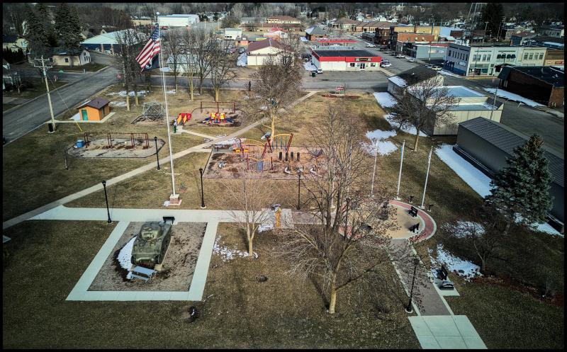 An aerial view of a small town park taken from about flag pole height. In addition to swings, slides, and the other usual playground equipment, this park has an old tank in it.