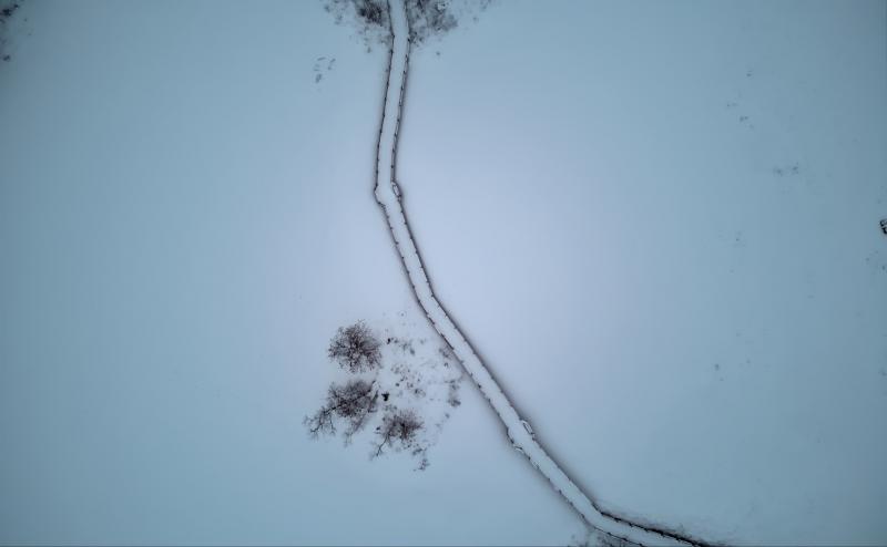 Drone photo from about 300 feet up, looking directly down at walkway across a snow covered lake. Walkway bends as it crosses the lake and a small island, with a few small trees is visible about half way across. 