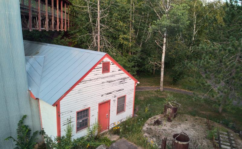 White single story building with red trim and aluminum roof attached to larger aluminum structure. Some kind of catwalk structure protrudes over white building into the surround forest. To the right, what looks like an old iron fire grate.