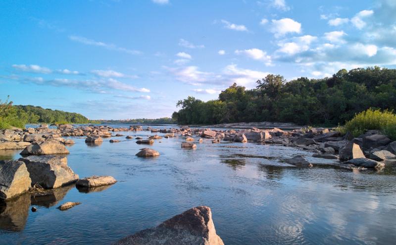 Photograph taken very close to the water looking downriver through an area dense with rocks sticking up out of the water. Tree line the banks on either side of the river and the sky is blue with scattered clouds.