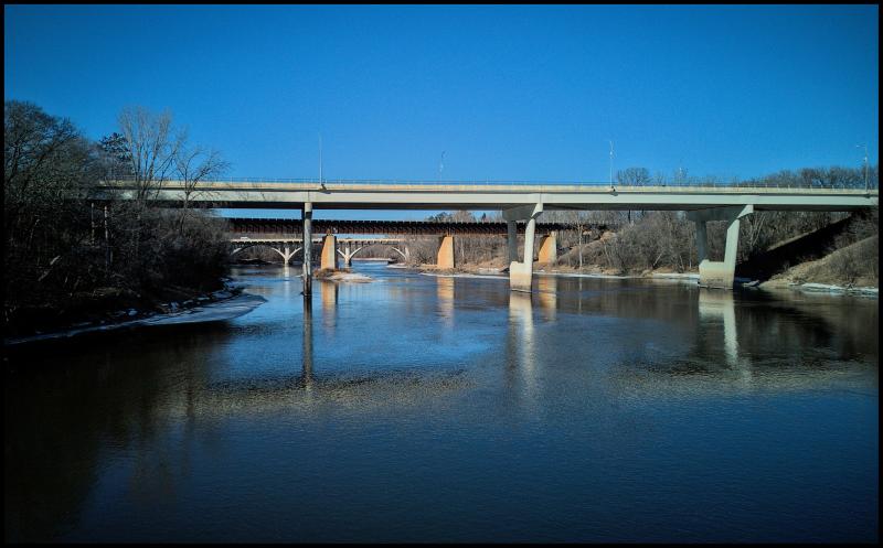 View of a bridge over the Mississippi River. We can see two additional bridges in background as we look underneath the first bridge. There is minimal snow and ice on the banks of the river.