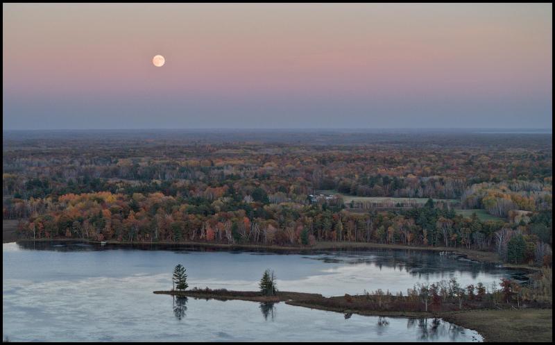 Aerial photo of a penisula sticking out into a calm lake just after sunset. The trees on the banks of the lake are showing their autumn colors and a full moon is visible in the sky.