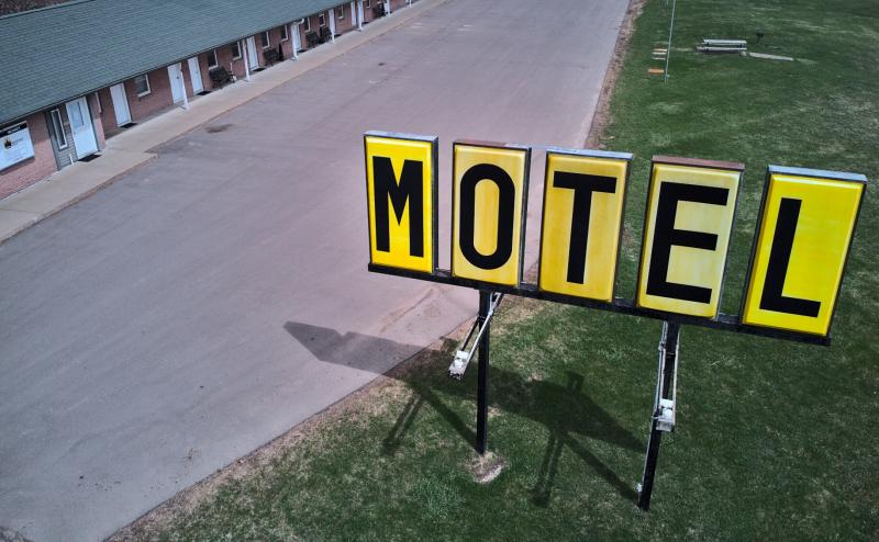 Aerial photo of a yellow motel sign with each letter in large block letters. Background is a glimpse of the oldstyle single floor motel, with doors to each room facing out to the highway. 