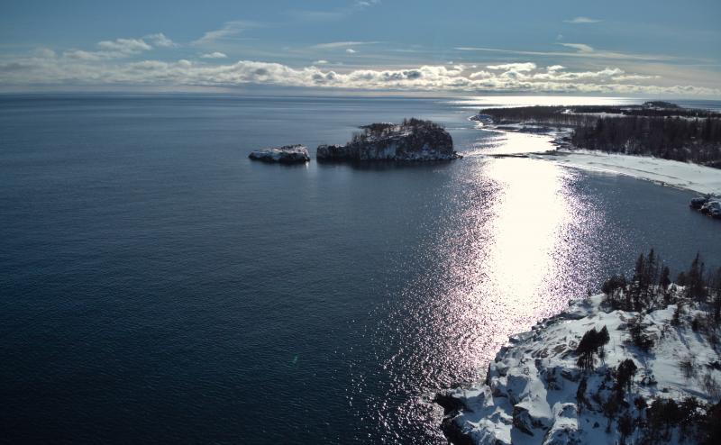 Drone photo of the shores of Lake Superior with several rock islands. Shores are covered with snow, but water is open and reflecting the sun.