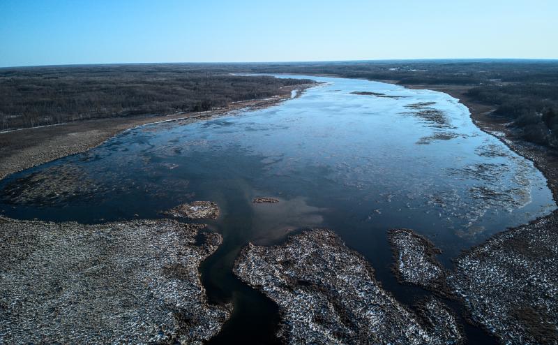 Aerial photo of Lake Ogechie near Mille Lacs Lake where the Rum river begins. Wec can see stream flow into lake through a swamp with a light coad of snow. Lake is mostly open with some large chunks of ice in the center. 
