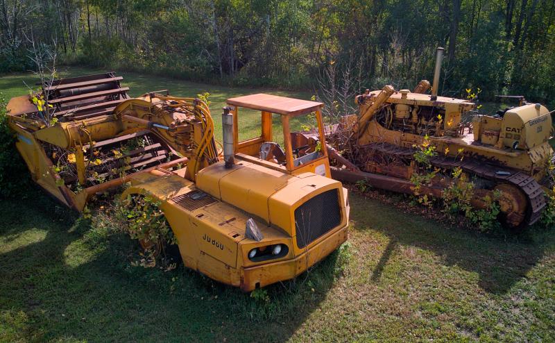Photo of bulldowser and another large piece of contruction equipment. Both items are yellow and overgrown with brush and weeds. Photo taken with drone at a downward angle from position a little higher than the equipment.