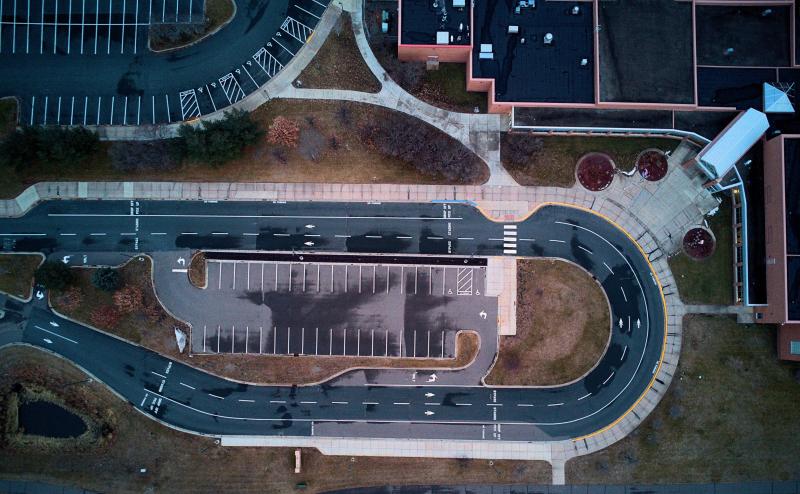 Drone photo looking straight down at small parking lot outside of school entrance. Two lanes of traffic circle the small parking lot with arrows directing traffic in one direction. Another parking lot nearby and parts of the school roof are also visible.