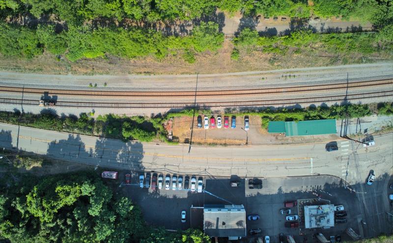 Looking straight down on a mix of things. From top to bottom, we see trees, bike path, train tracks cars in parking lot, road, more cars and buildings. 