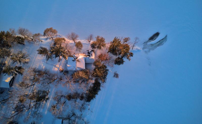 Looking straight down on a house/cabin on a snowy peninsula justting into a frozen lake. It's near sunset and the buildings and trees are casting interesting shadows.