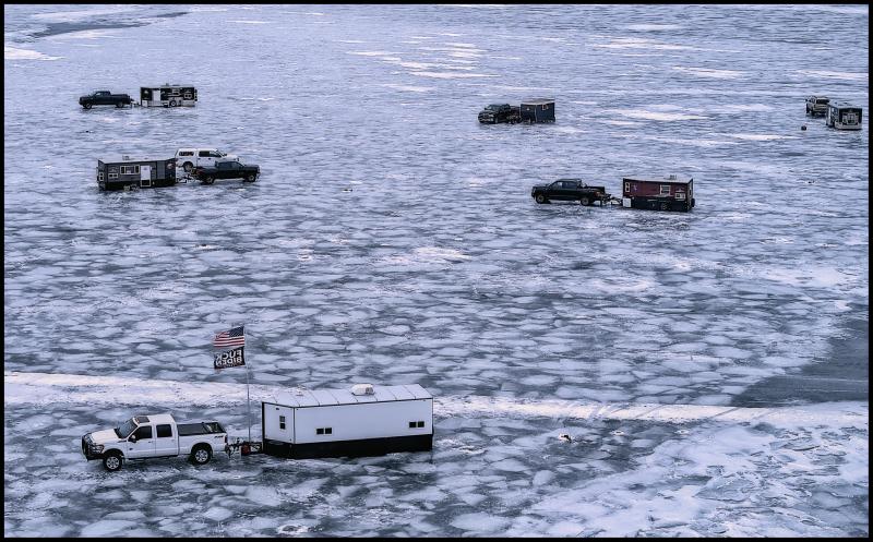 Aerial view of a half dozen 'pull behind' ice houses all attached to pick up trucks on a frozen lake. One can see lots of small chuncks of broken ice that have froze back together through the surface of the ice. The ice house in the foreground is flying an American flag along with one that say's "F--k Biden."