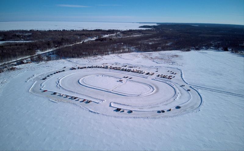 Drone photo from distance of a race track for cars plowed on a frozen lake. 50-100 cars are participating or spectating in races.