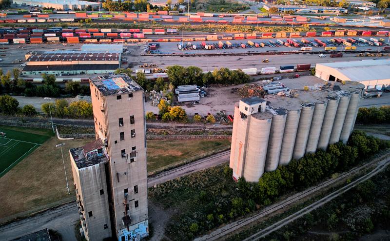 Aerial view of an abandoned grain elevator near an active rail yard. The rail yard has many rail cars light up slightly by the sunset happening behind the camera.