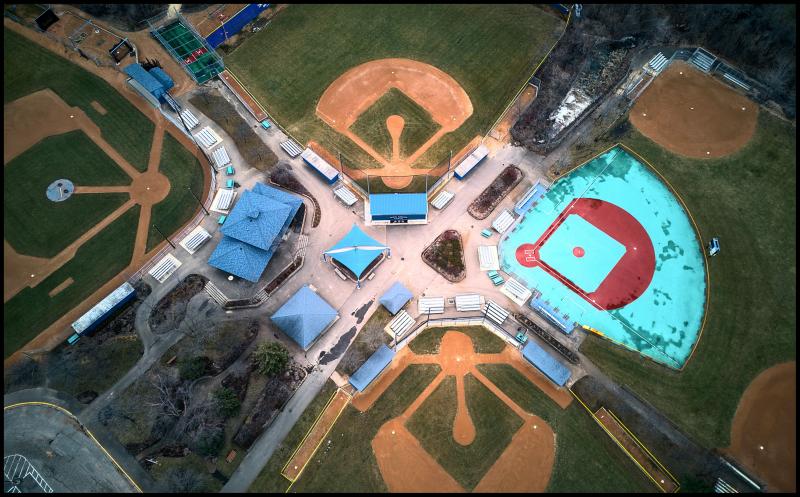 Aerial view looking down on complex of several baseball fields some small buildings for concessions and other stuff in the middle. One field is light blue, while the others are green.
