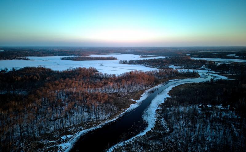 Aerial photo of an open river flowing into a frozen lake. It's near sunset and you can see the sun in the trees. The woods on either side of river are covered in scattered snow. 