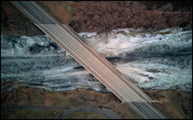 Aerial view, looking straight down on a frozen river, horizontal in the image. A bridge cuts through the image and across the river at an angle. The frozen surface of the river is very different colors and textures.