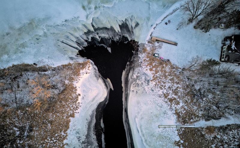 Looking straight down at water flowing from ice covered lake into an open stream or small river. Jagged ice formations form the teeth in the mouth of the river.